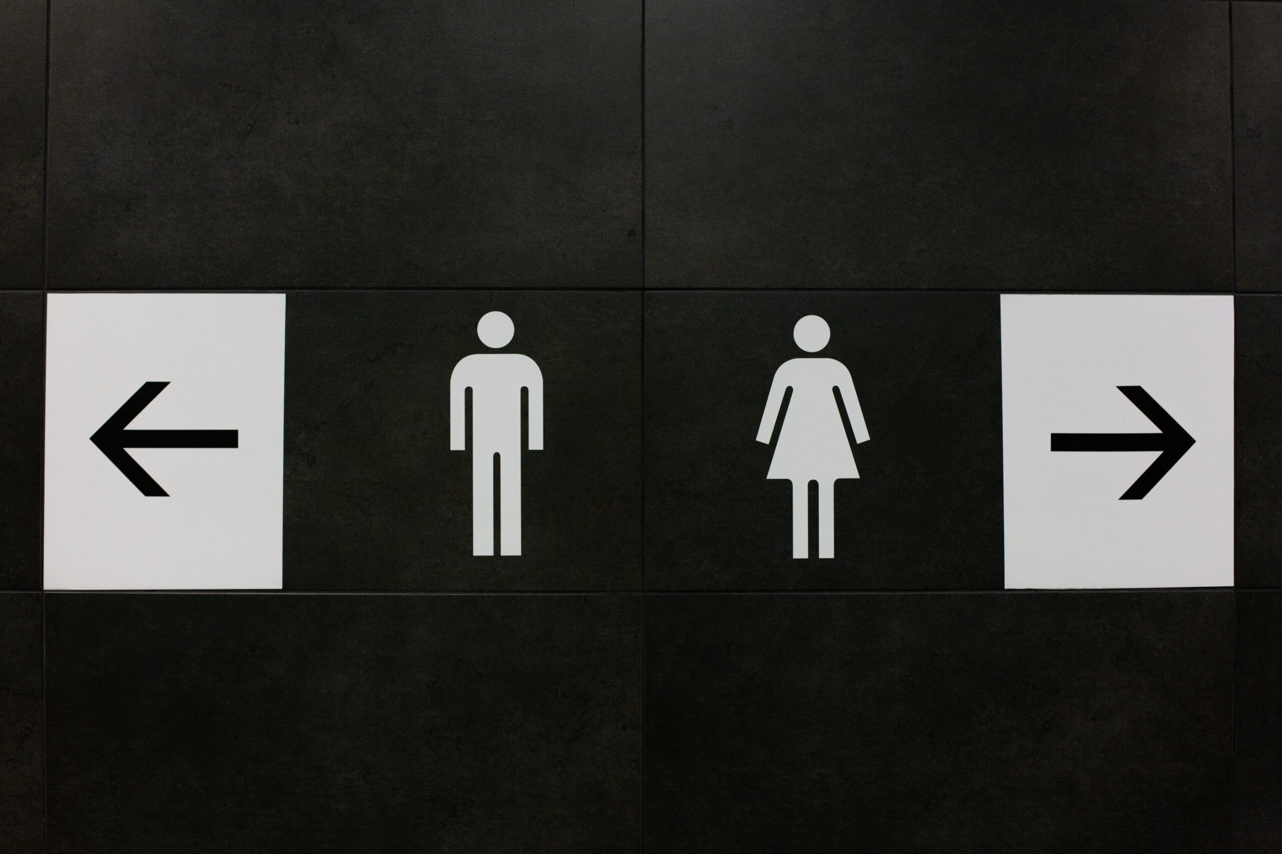 Black and white "Men's" and "Women's" bathroom signs, flanked by arrows pointing right and left.064435-scaled Black and white "Men's" and "Women's" bathroom signs, flanked by arrows pointing right and left.
